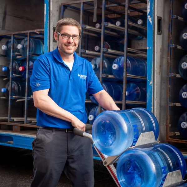 water delivery driver standing in front of truck, delivering 4 water cooler bottles and a case of water