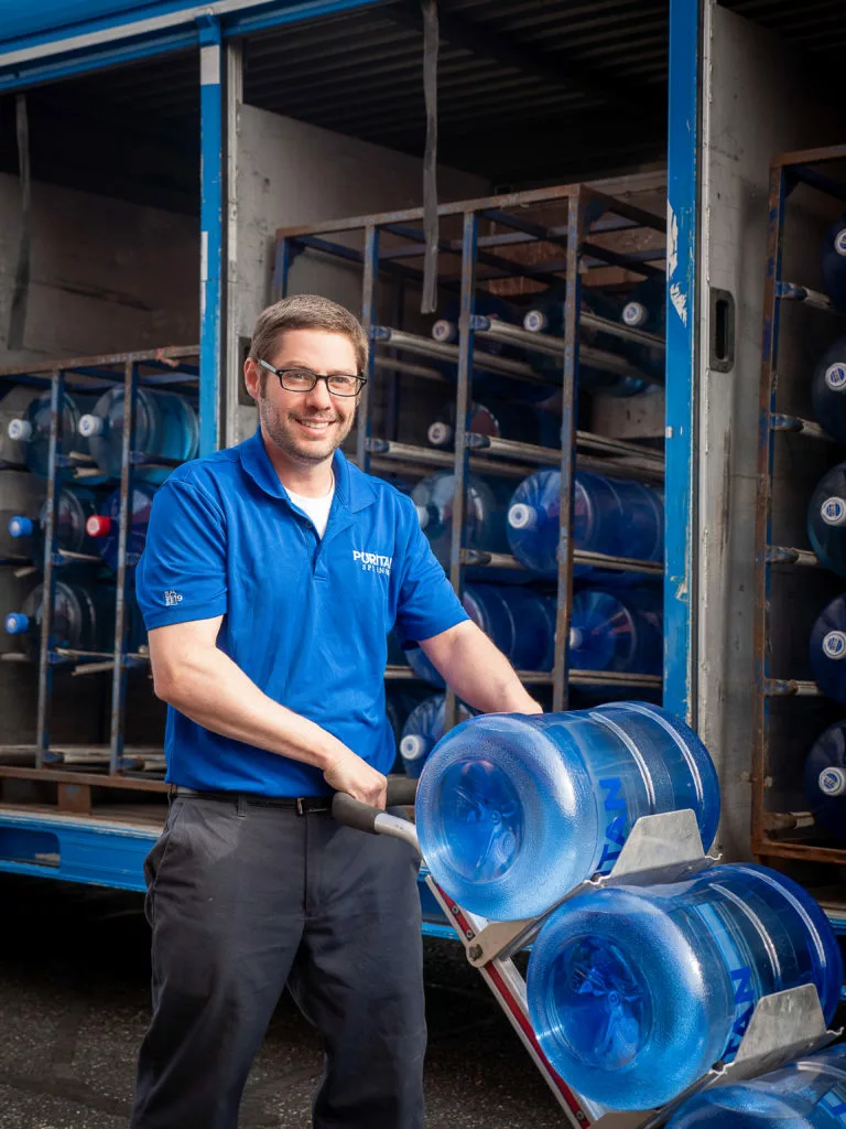 water delivery driver standing in front of truck, delivering 4 water cooler bottles and a case of water